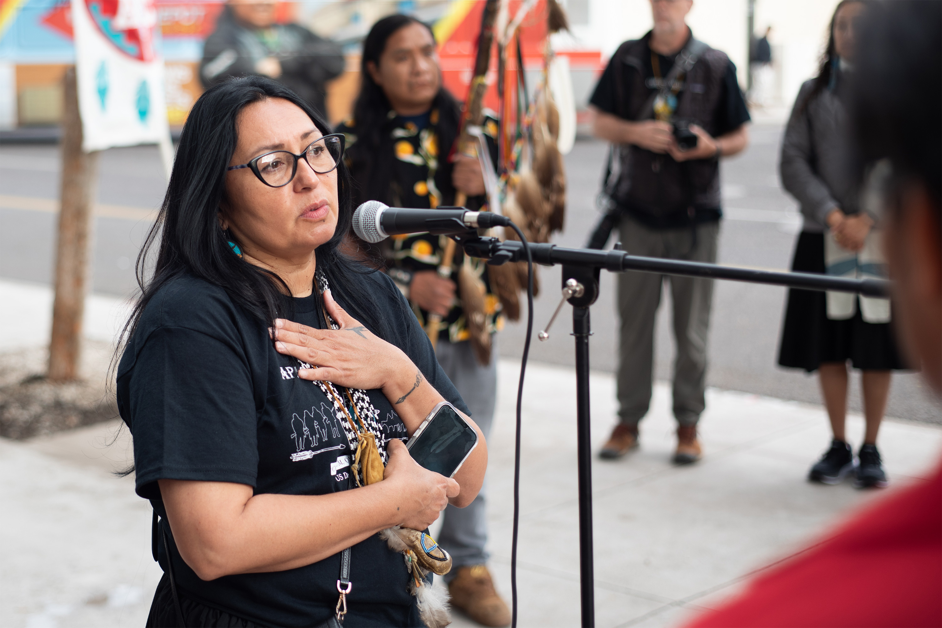 A photo of an indigenous woman speaking into a microphone at an event in front of a courthouse.
