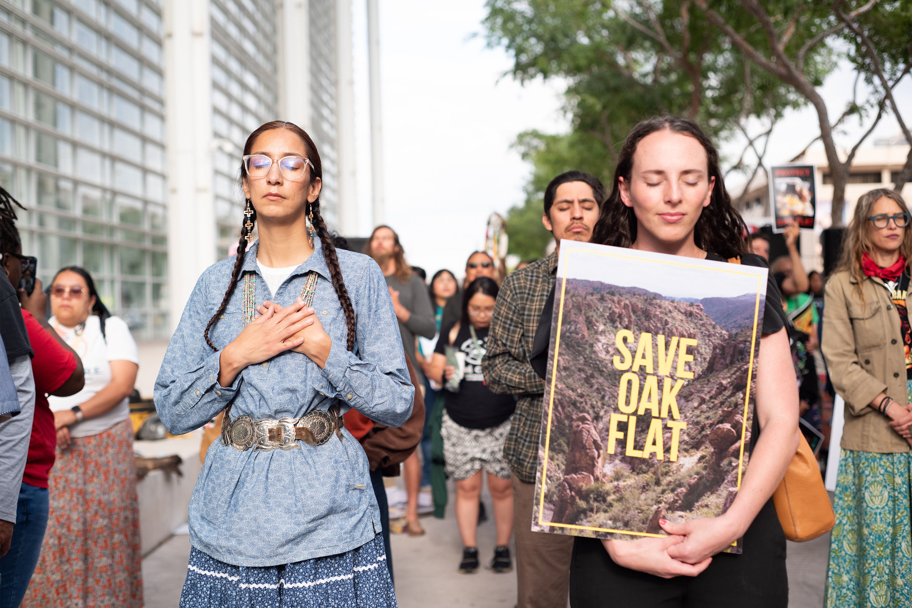A photo of two women bowing their heads in prayer. The woman on the right holds a sign that reads, "Save Oak Flat."