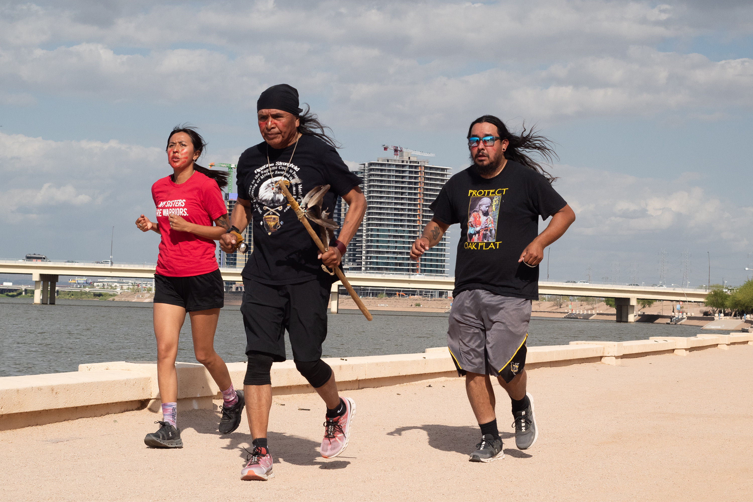 A photo of three runners of indigenous descent running along a sidewalk. They wear garb with protest slogans on them.