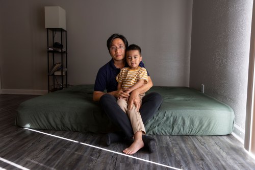A young boy sits on his father's lap on a mattress covered in a green fitted sheet on the floor.