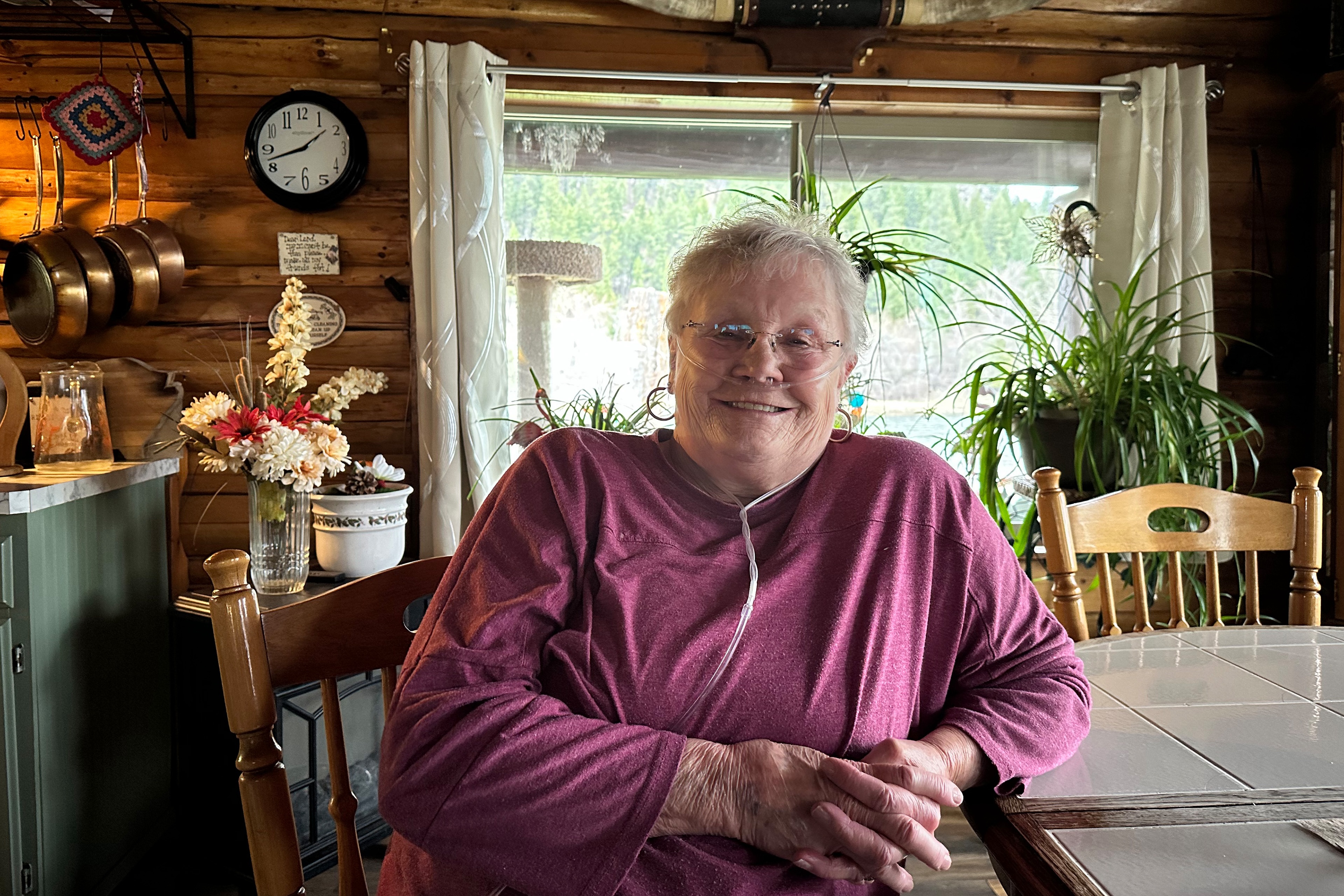 A woman in a maroon longsleeve shirt and glasses, sits at a table with her hands folded. A portable oxygen machine is helping her breathe