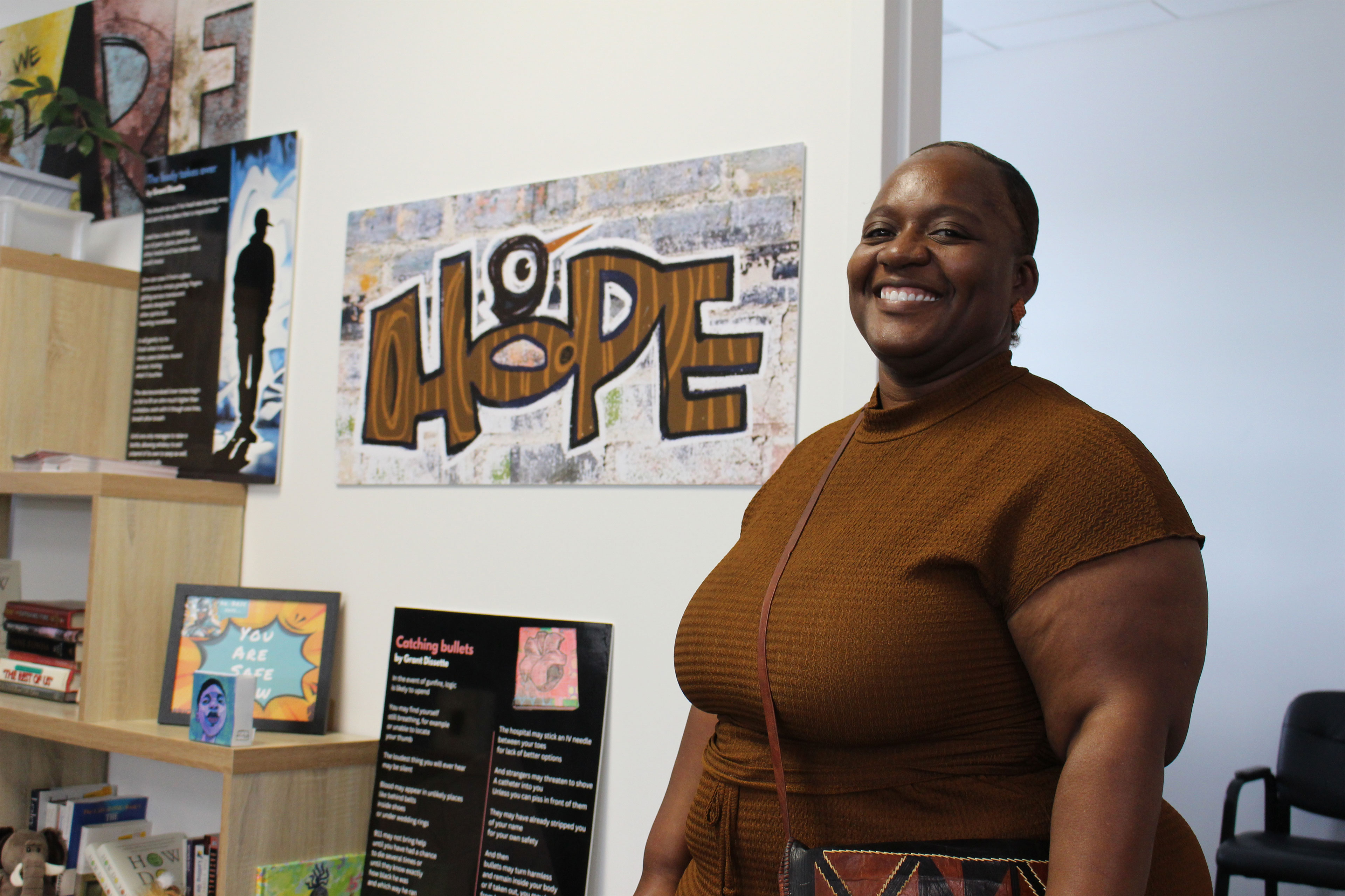 A woman wearing dark brown stands and smiles at the camera. There are several powers on the wall behind her and the most prominent of them reads "Hope."