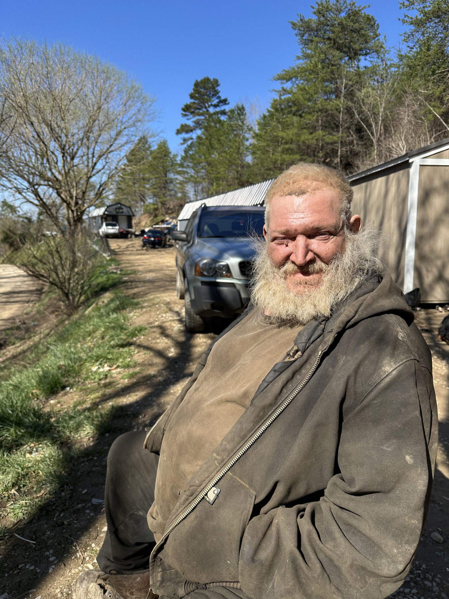 A man with a blonde beard and wearing a tan shirt and zippered sweatshirt sits outside and smiles at the camera.