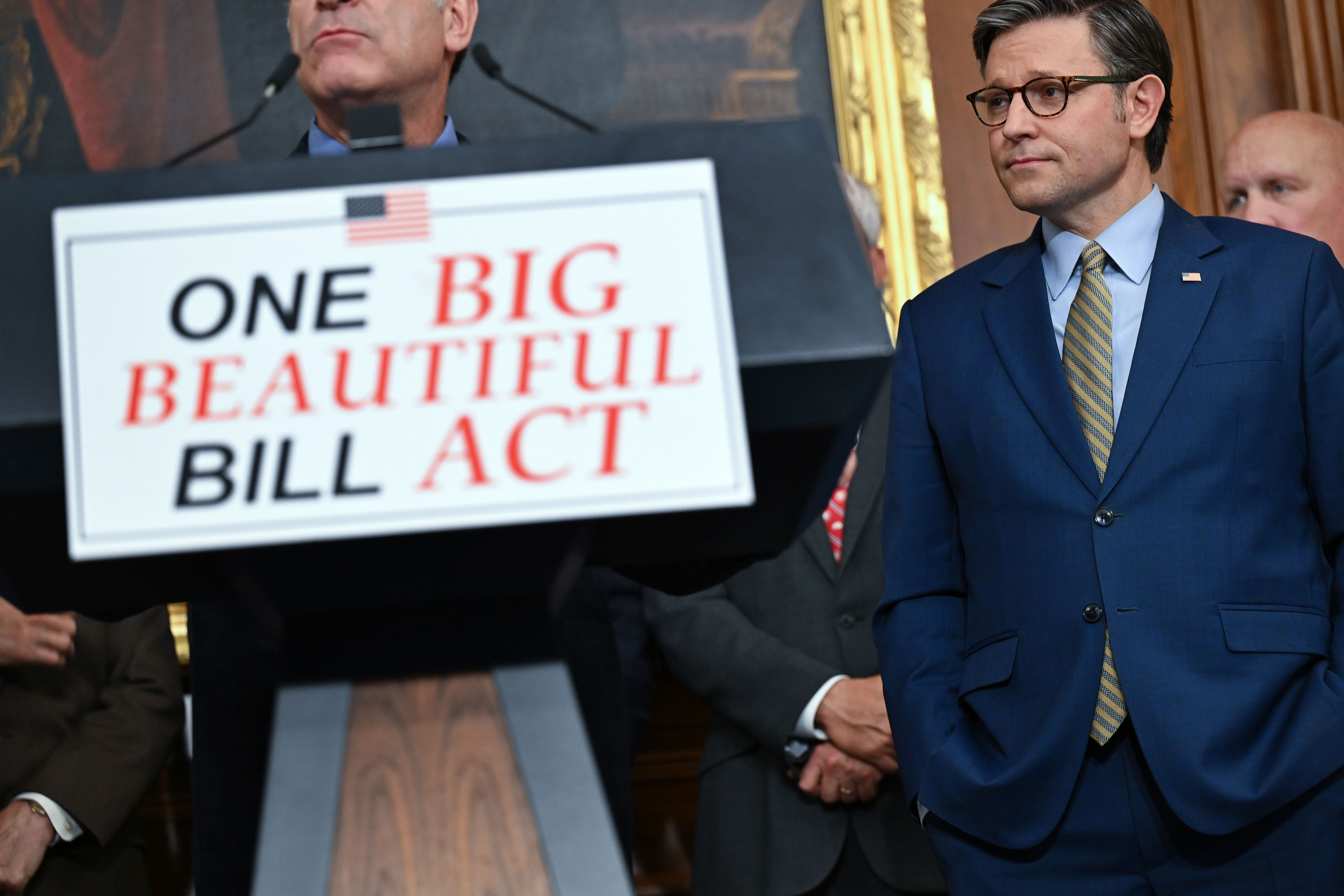 A close-up photo of a lecture with a sign reading "One Big Beautiful Bill Act" on its front. A person stands behind the lecture, but the top half of his face is out of the shot. Speaker of the House Mike Johnson is seen standing to the right of the lecture.