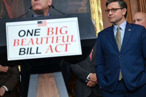 A close-up photo of a lecture with a sign reading "One Big Beautiful Bill Act" on its front. A person stands behind the lecture, but the top half of his face is out of the shot. Speaker of the House Mike Johnson is seen standing to the right of the lecture.