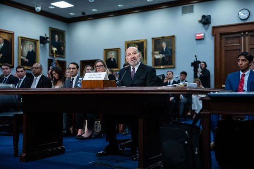 A balding man with a gray beard sits behind a table and speaks into a microphone. He wears a dark suit and purple tie and a sign on the table reads "The Honorable Howard Lutnick." People sit in several rows of chairs behind him.