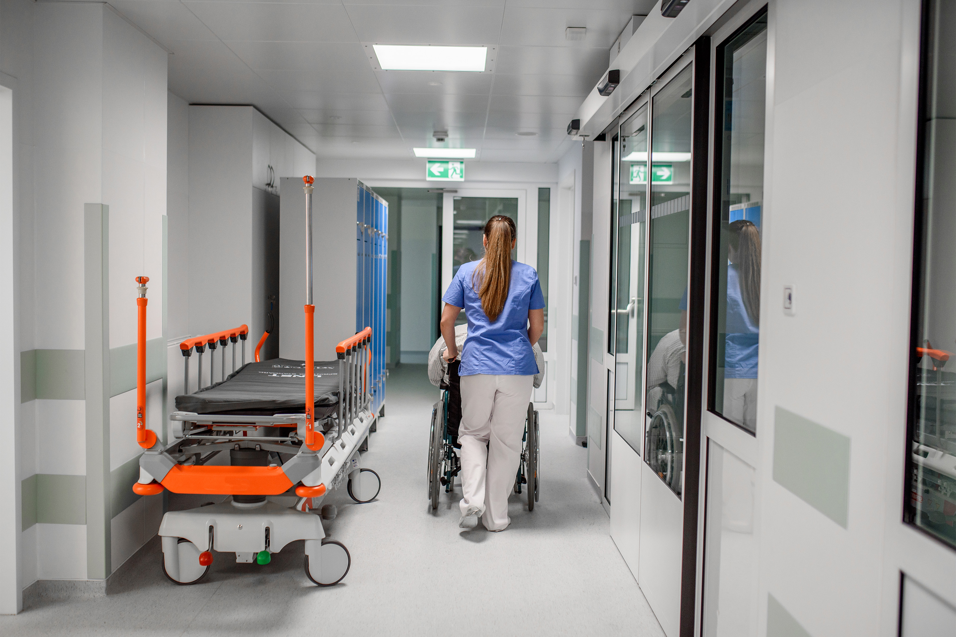 A photo of a nurse pushing a patient in a wheelchair in a hospital corridor.