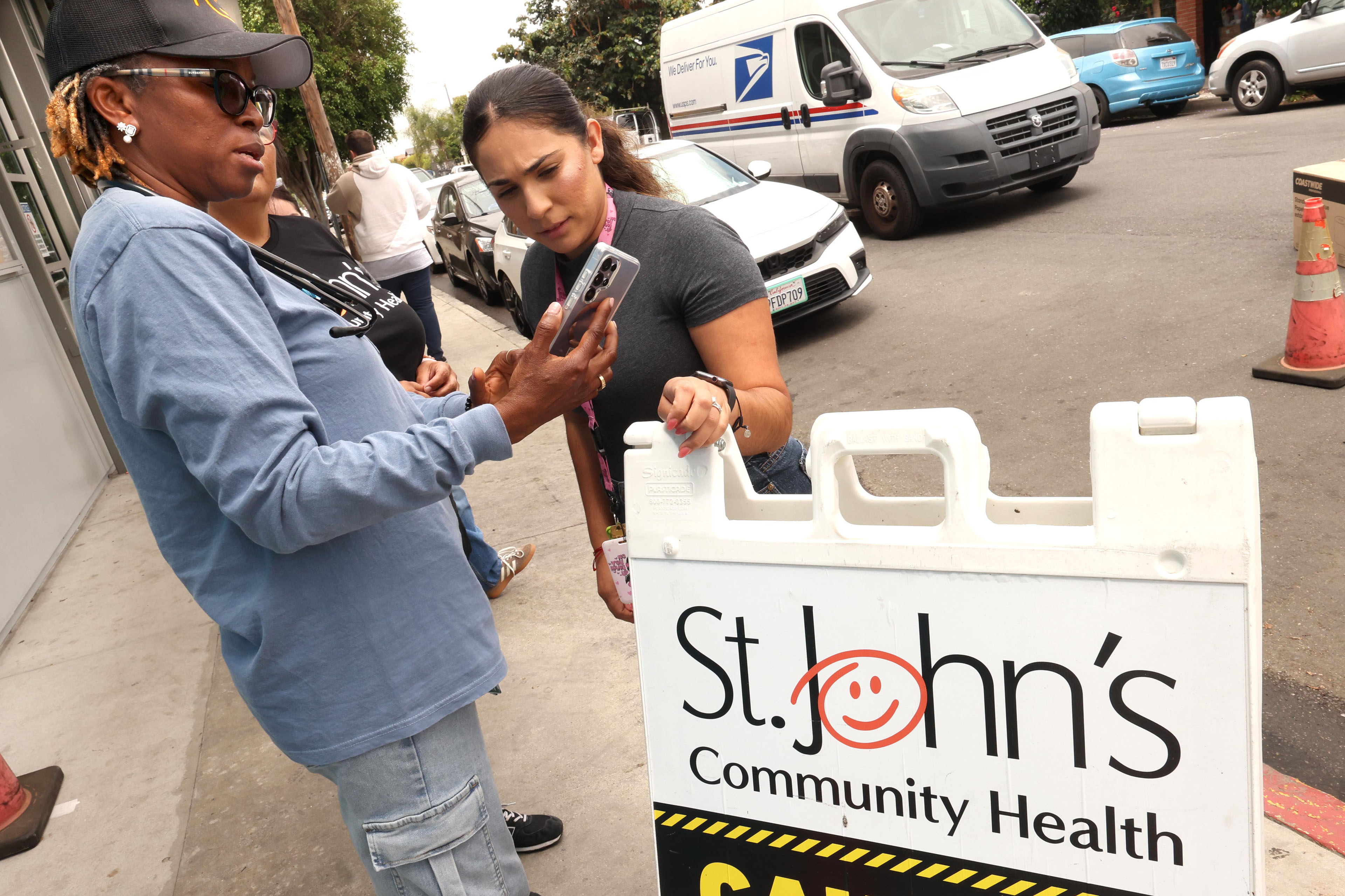 A woman is showing another woman something on a cell phone screen. In front of them is a sign that reads, "St. John's Community Health."