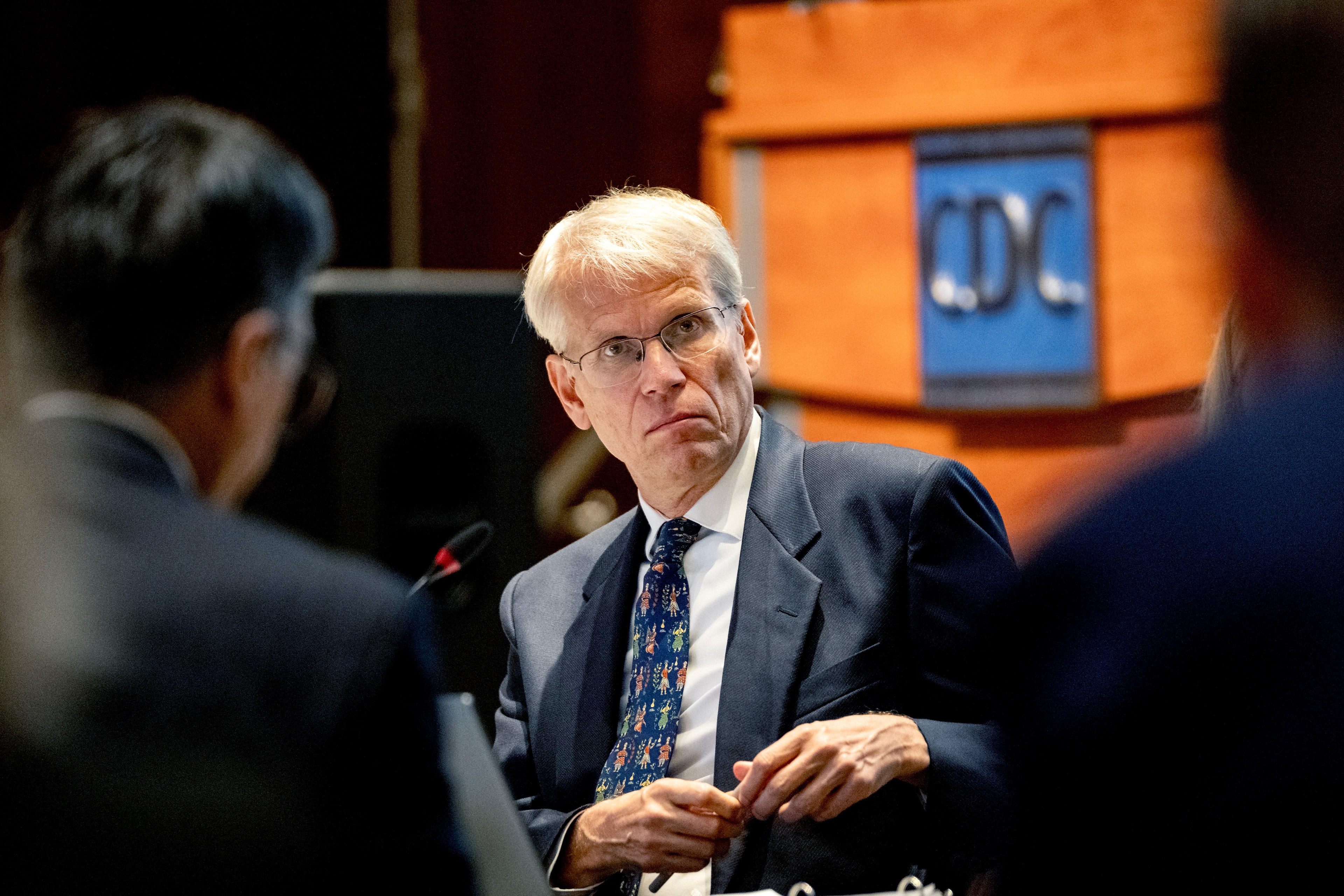 An older man in a suit and tie with glasses is seated while a sign that reads "CDC" is in the distance behind him