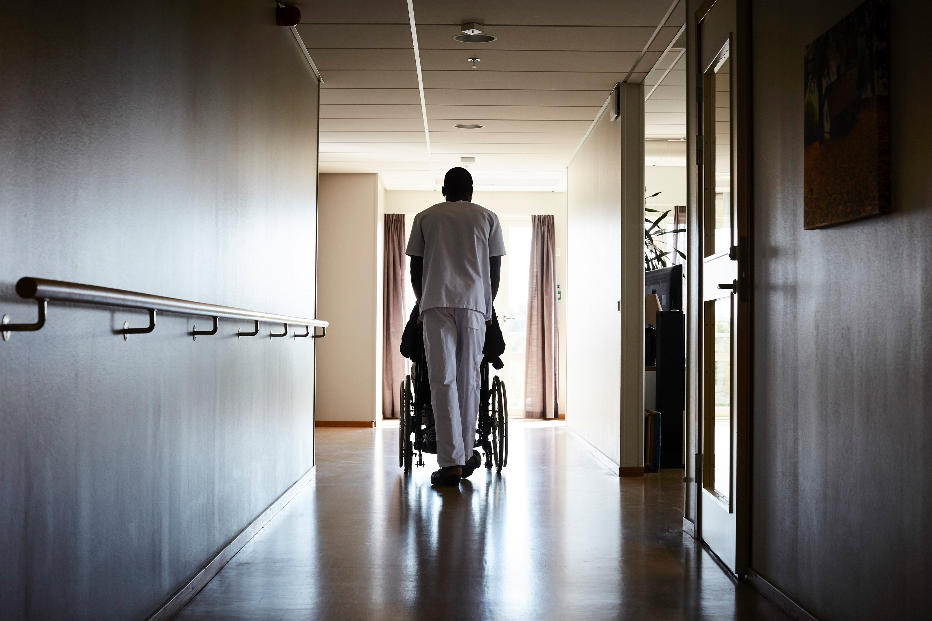 A photo of a nurse wheeling his patient in a wheelchair in a nursing home.