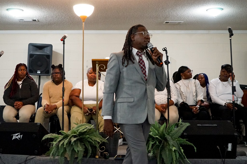 A pastor, wearing a grey suit and gingham tie, holds a microphone as he speaks to an audience.