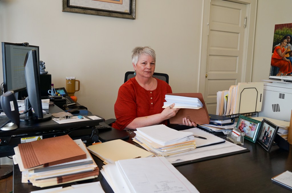 A woman sits at a desk covered in stacks of paper and two computer monitors.