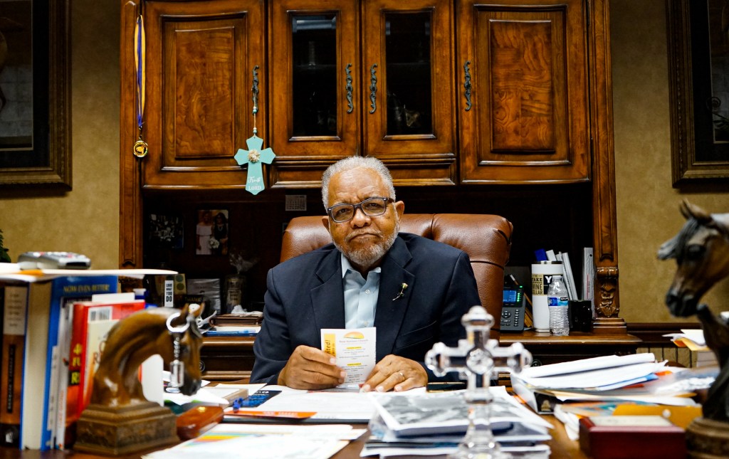 A man faces the camera as he sits his desk that is covered in papers and books.