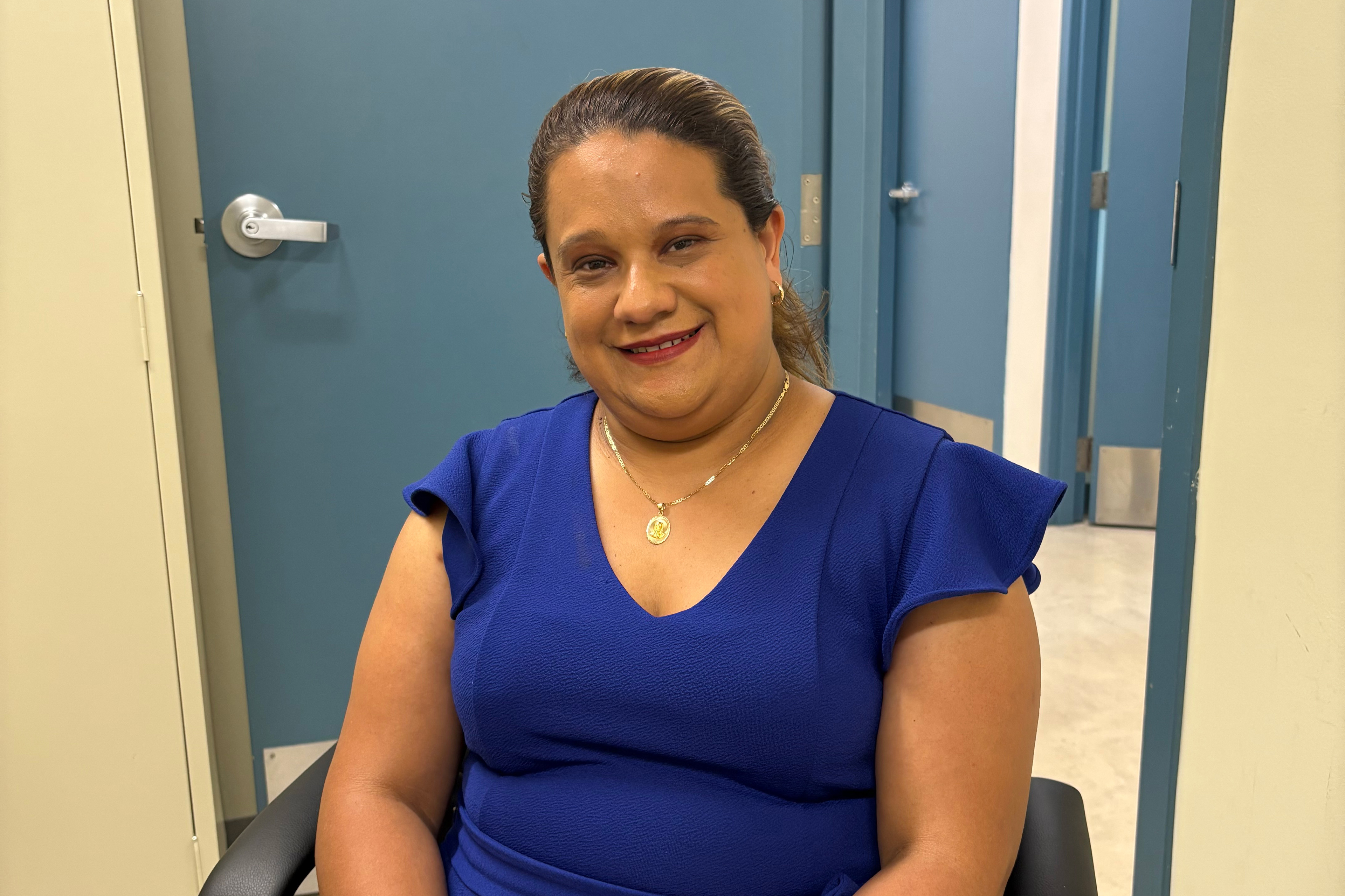 A woman in a blue tshirt with dark hair and red lipstick smiles at the camera. She is seated in front of a blue door