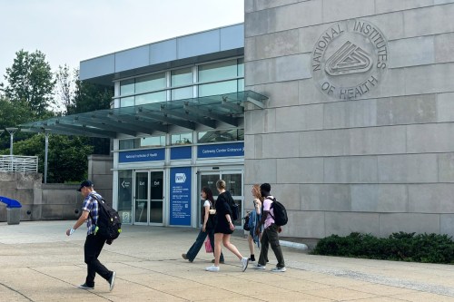 A photo of the NIH campus on a partly-cloudy day. A few pedestrians walk past an entrance to the building.