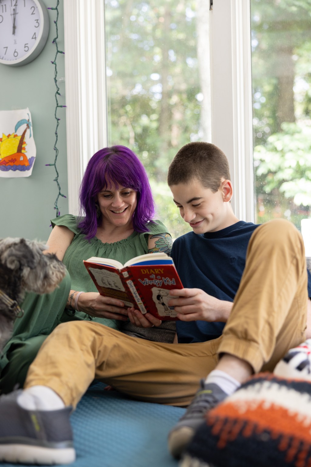 A woman with purple hair and her teenage son, who is wearing mustard colored pants, sit on a blue couch, reading a book.