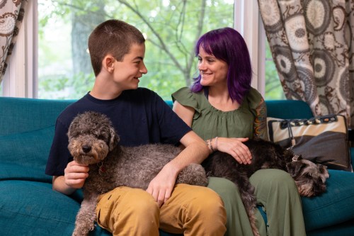 A teenage boy and his mother sit on a teal couch, side by side, smiling at one another. A dog is seated on each of their laps.