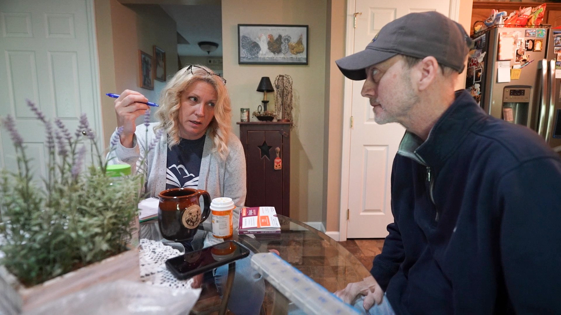 A photo of a woman speaking to her husband at the kitchen table. She is writing in a notebook and holding a blue pen. Her husband has a pill bottle and organizer in front of him.
