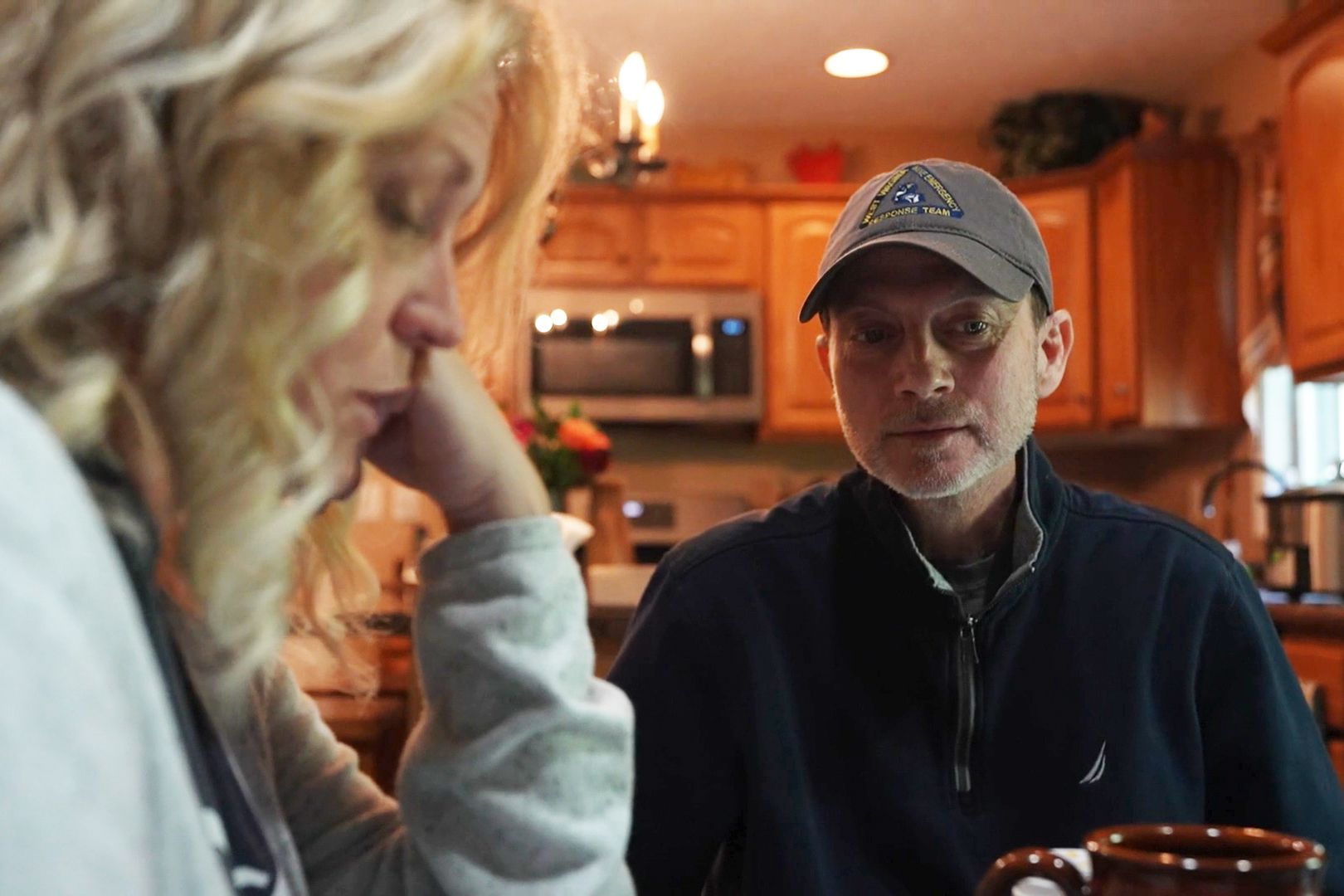 A husband sits at the kitchen table. His wife is seen close on the left of the frame. She is blurred slightly as the camera is focused on the husband.