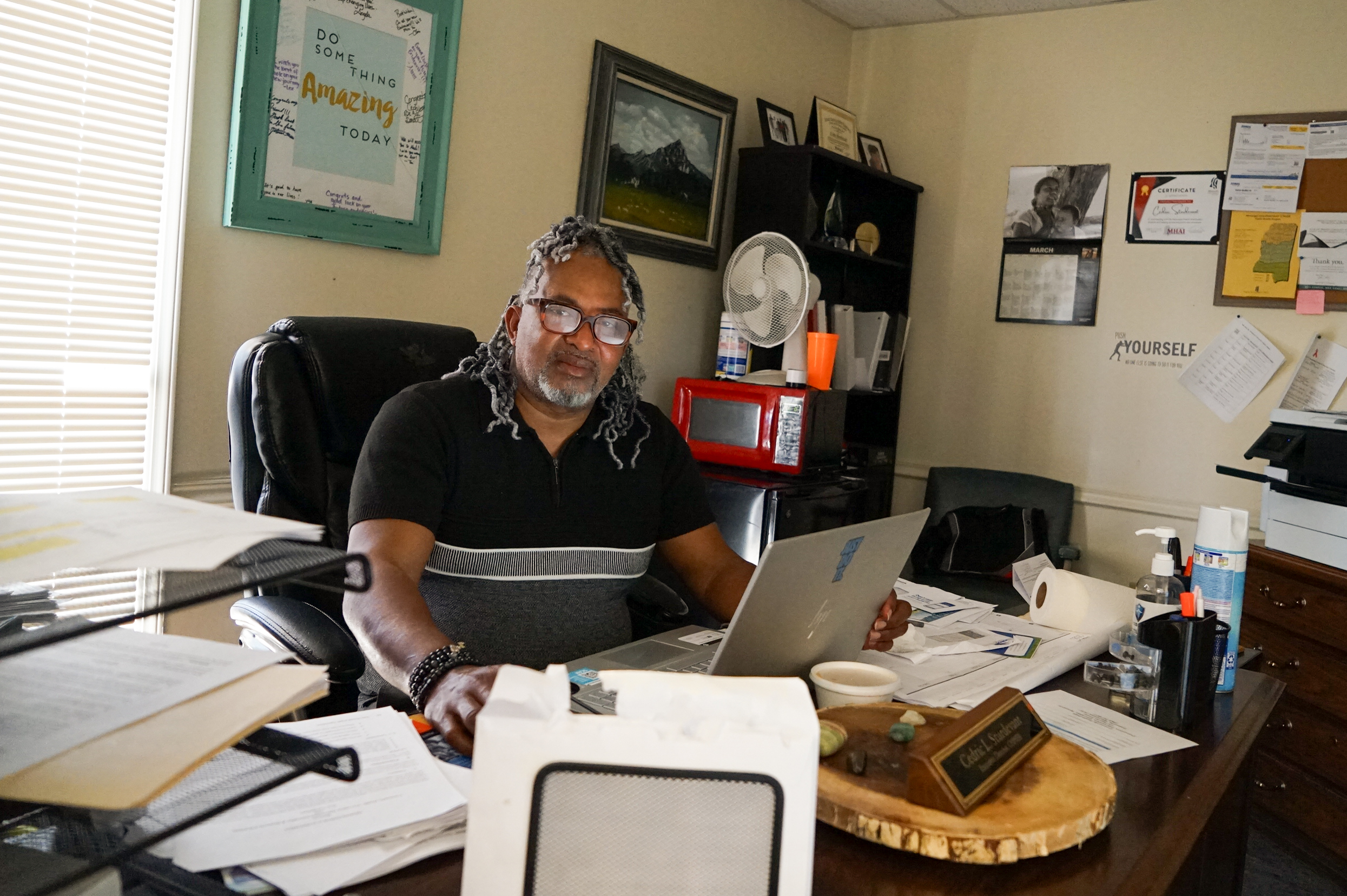 A man sits at an office desk that shows lots of signs of activity; stacks of paper, an open computer, and a name plate.