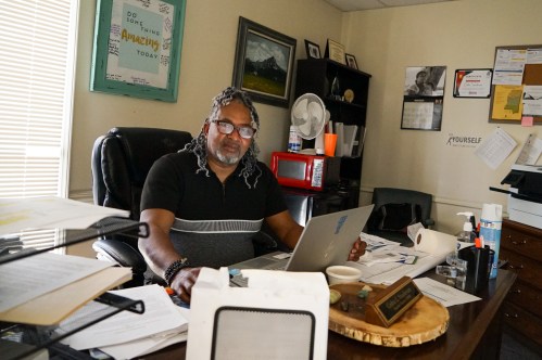 A man sits at an office desk that shows lots of signs of activity; stacks of paper, an open computer, and a name plate.