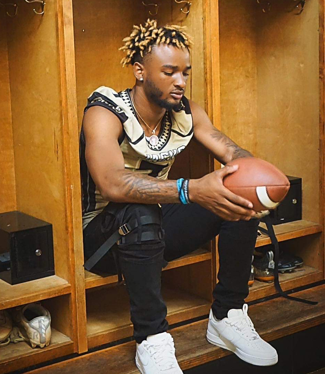 A photo of a young Black man sitting in a football locker room. He is wearing some of the gear and holding a football, looking down at it.