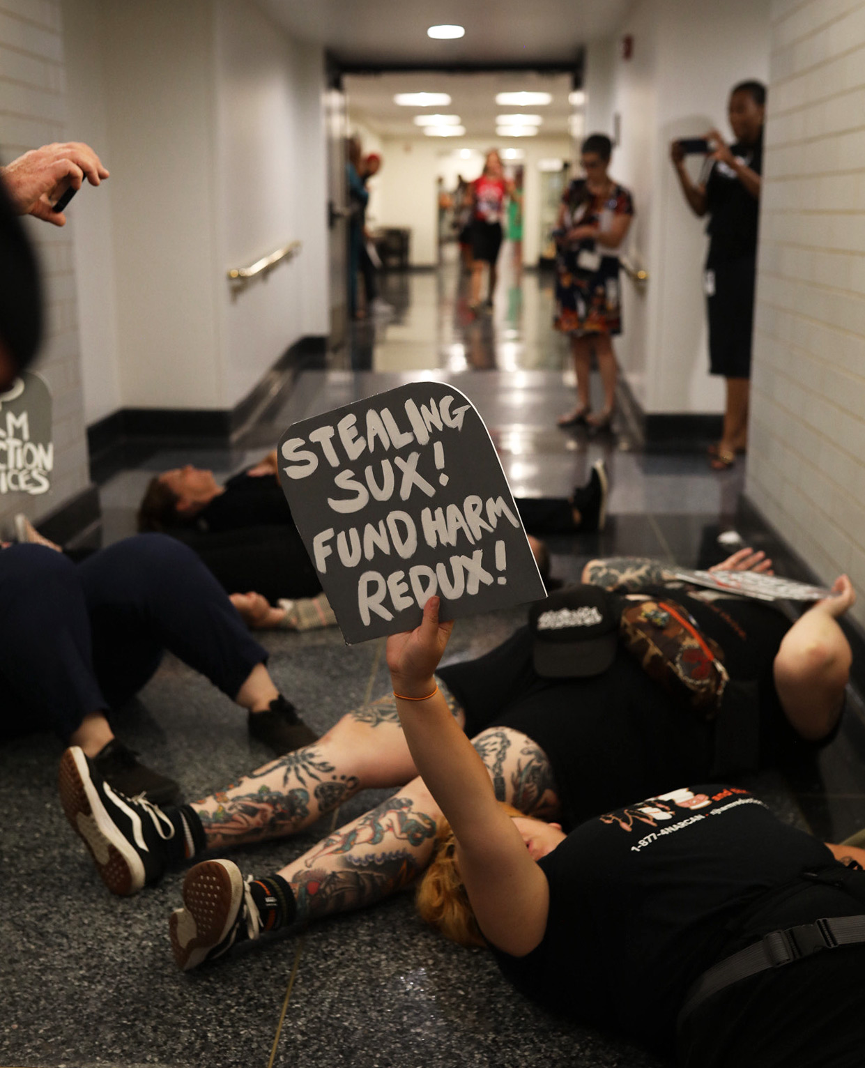 A photo of a die-in at the New Jersey Statehouse. One protester lying on the floor holds up a sign that reads, "Stealing sux! Fund harm redux!"