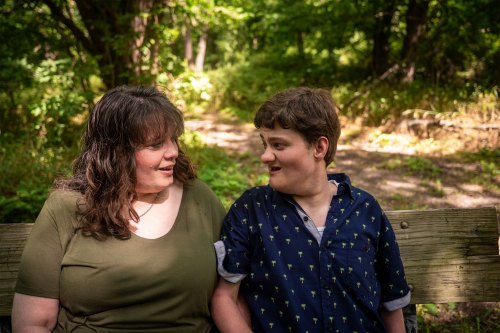 A woman with brown hair and wearing an olive green t-shirt sits on a bench and looks at the young man with brown hair and a wearing a blue shirt with small palm trees sitting on the bench beside her. They are in a wooded area and there are trees in the background.