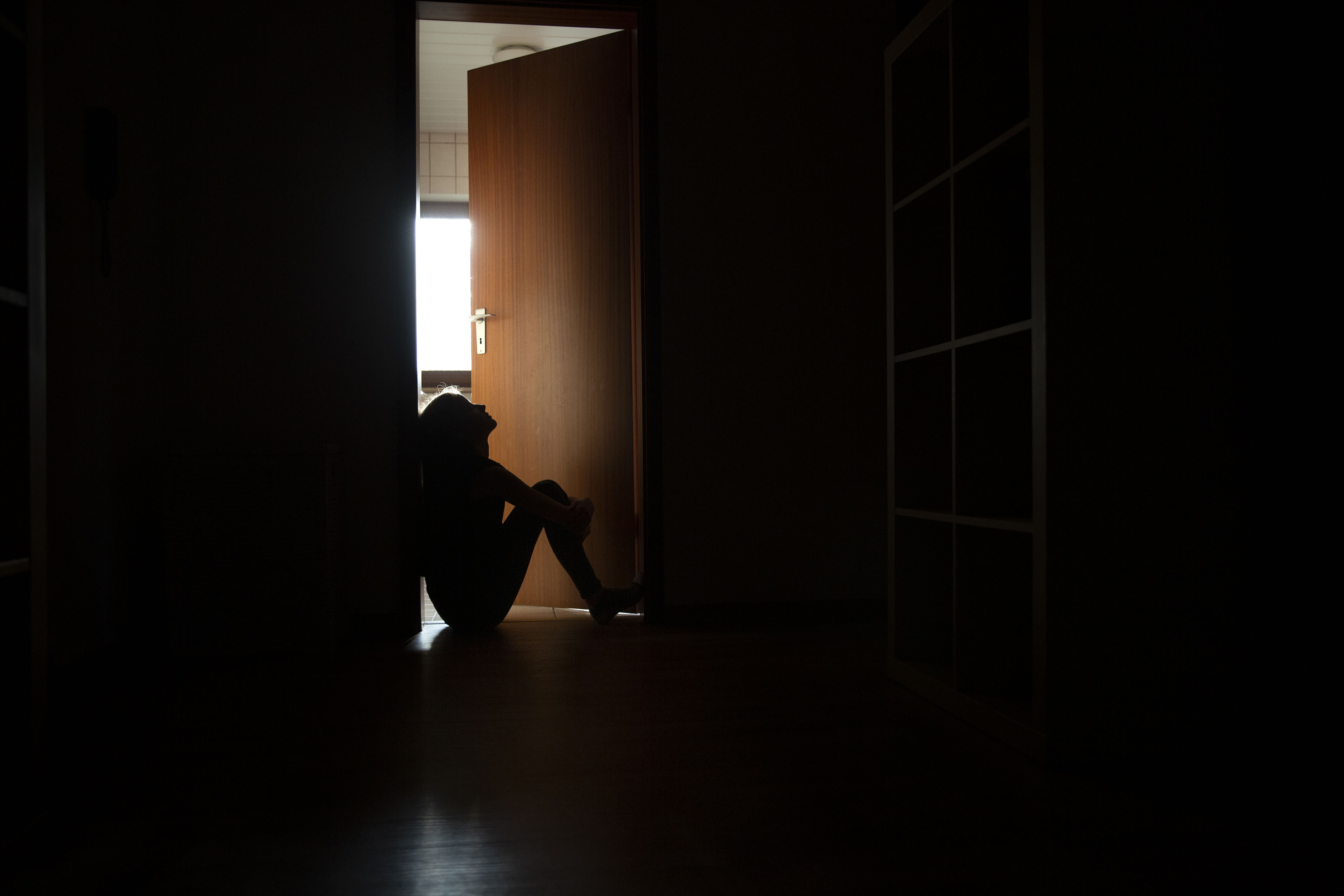 A photo of a teenager sitting in the frame of a doorway backlit by a room filled with daylight.