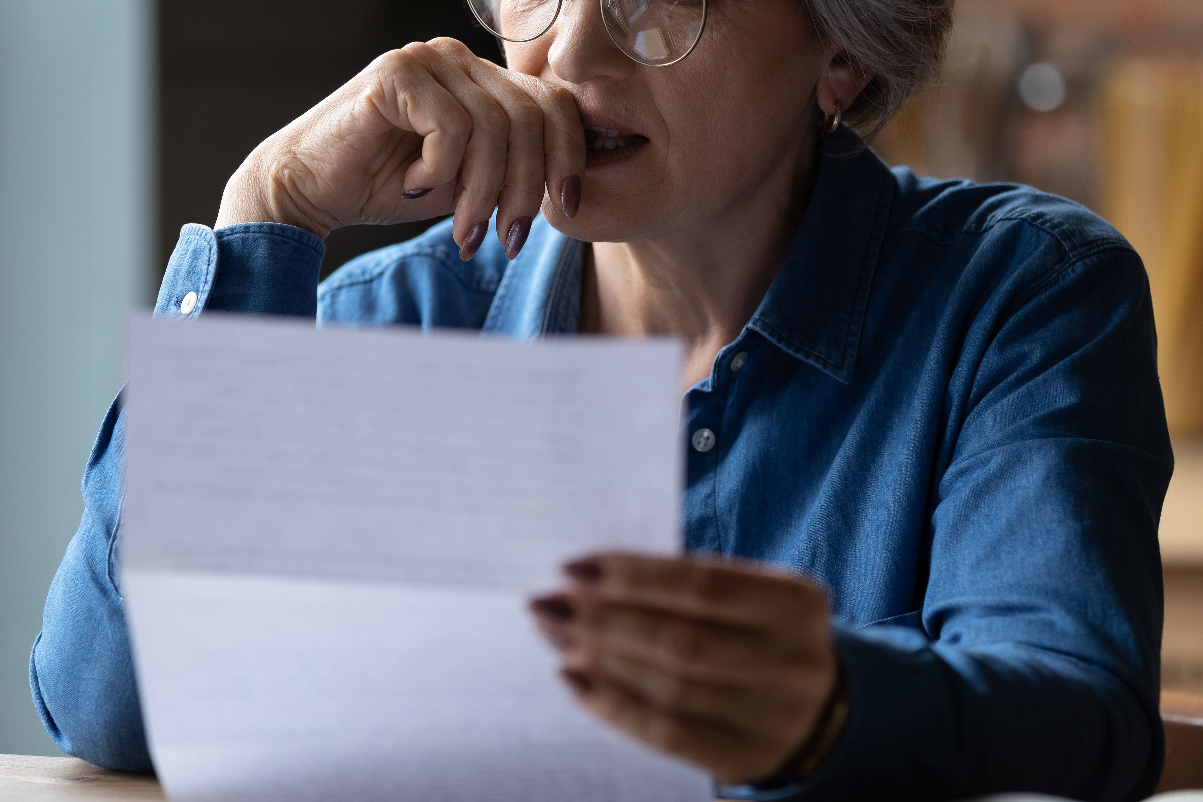 A closely cropped photo of a senior woman holding a paper letter. She presses her hand to her lips as she makes a stressed expression.