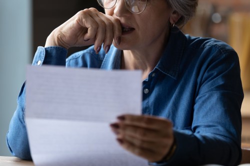 A closely cropped photo of a senior woman holding a paper letter. She presses her hand to her lips as she makes a stressed expression.