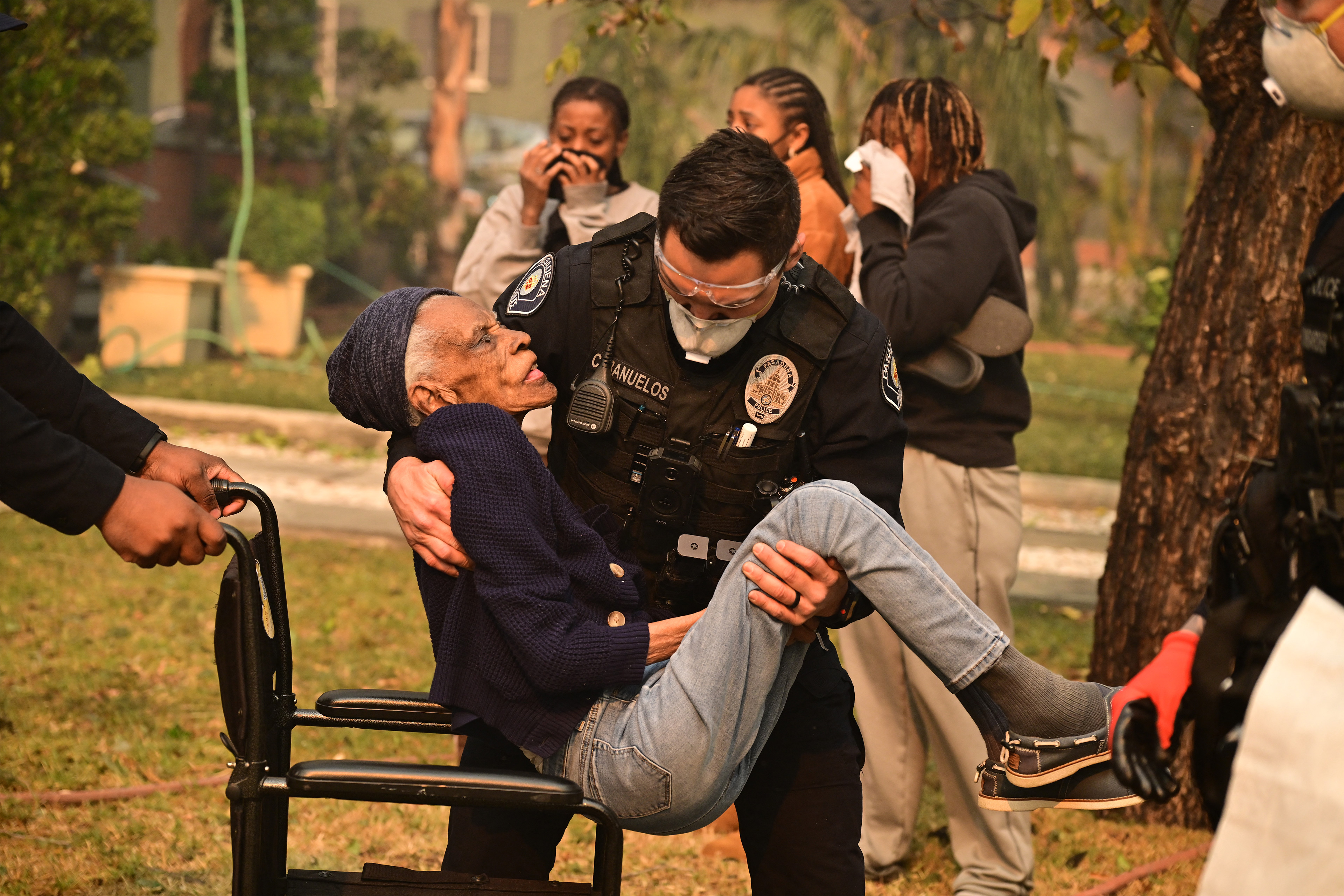 A photo of a police officer carrying an older woman. He is lowering her into a wheelchair.