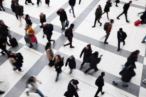 A photo of a train station as commuters and pedestrians walk hurriedly around the frame. Their forms are blurred as they are in motion.