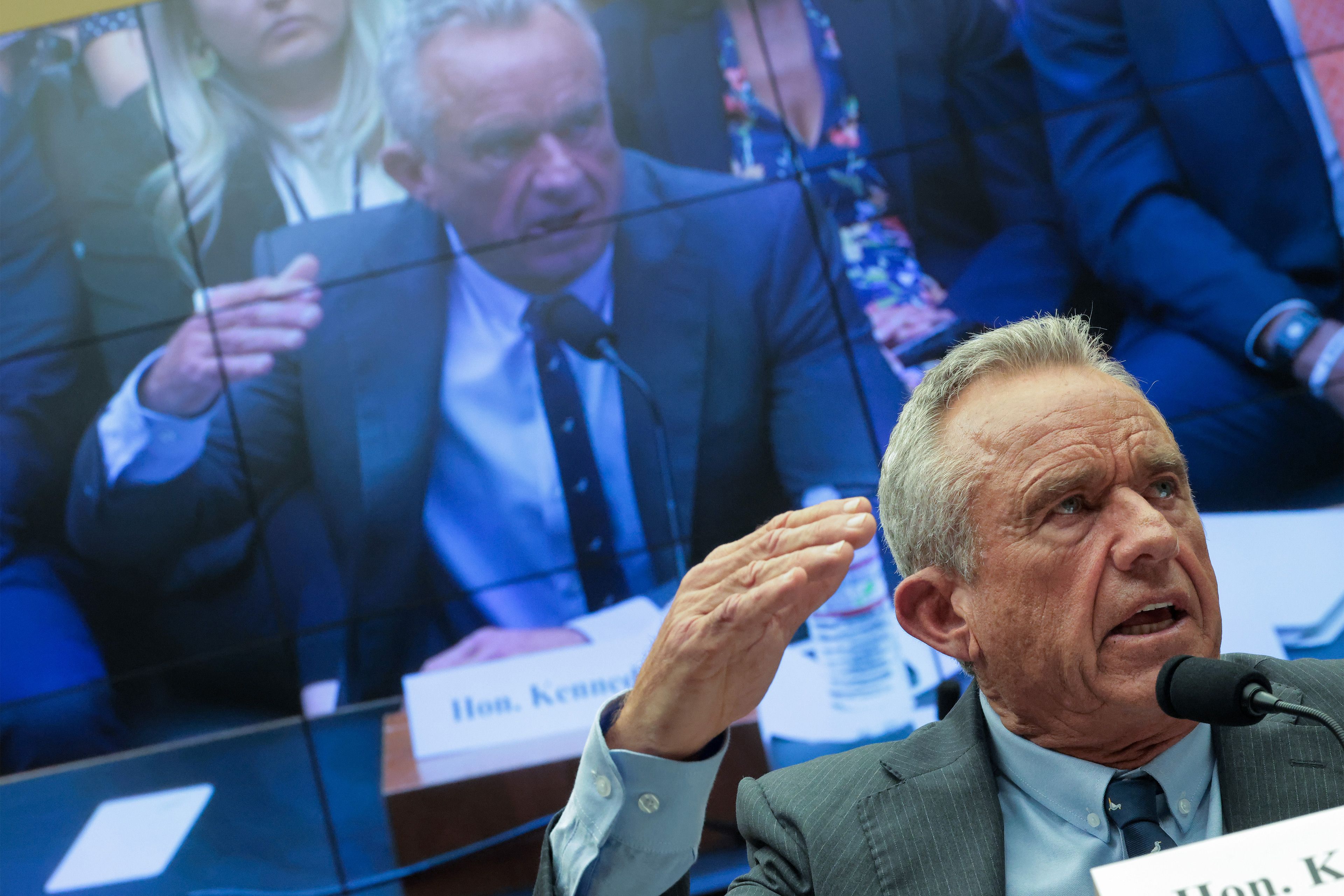 Health and Human Services Secretary Robert F. Kennedy Jr. speaking in a House of Representatives hearing room. He is also seen on a screen behind him broadcasting the hearing.