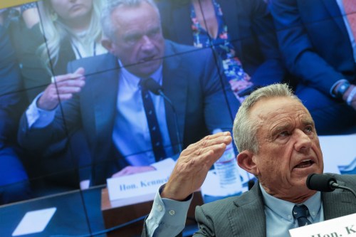 Health and Human Services Secretary Robert F. Kennedy Jr. speaking in a House of Representatives hearing room. He is also seen on a screen behind him broadcasting the hearing.