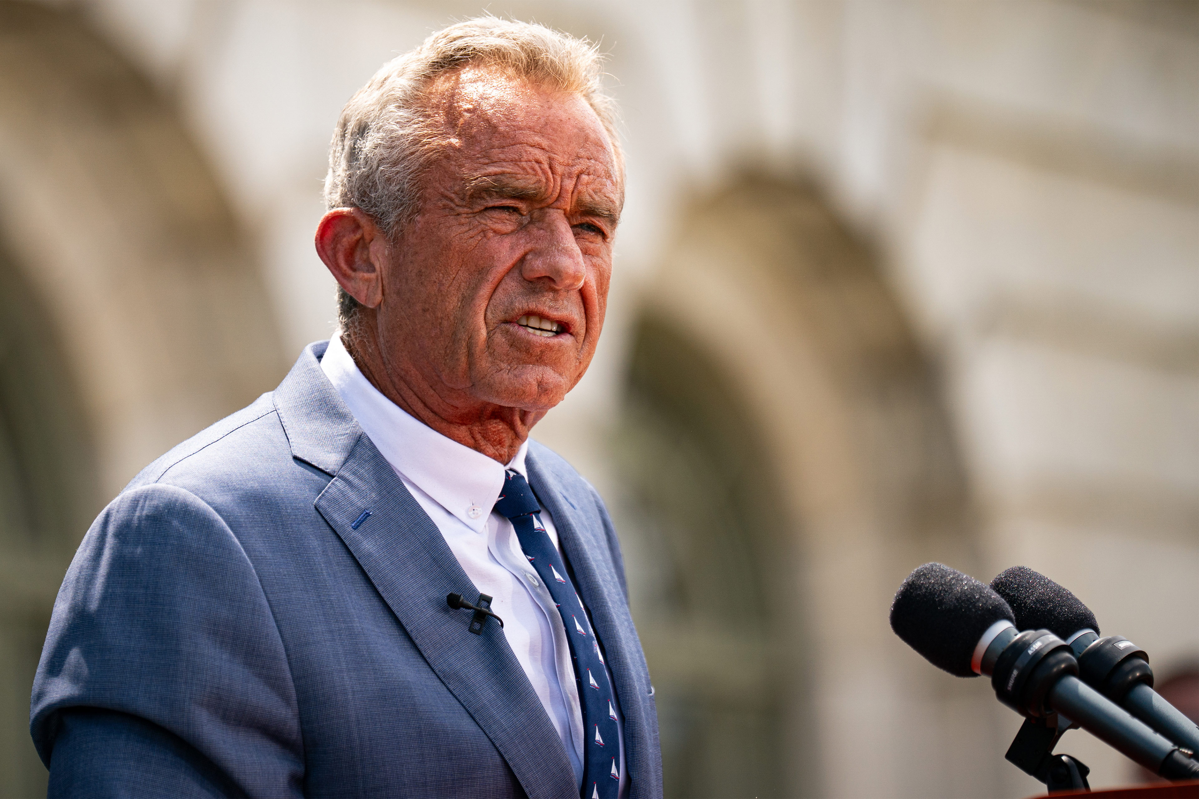 A photo of Robert F. Kennedy Jr. speaking in front of a building. Two microphones are seen next to him.