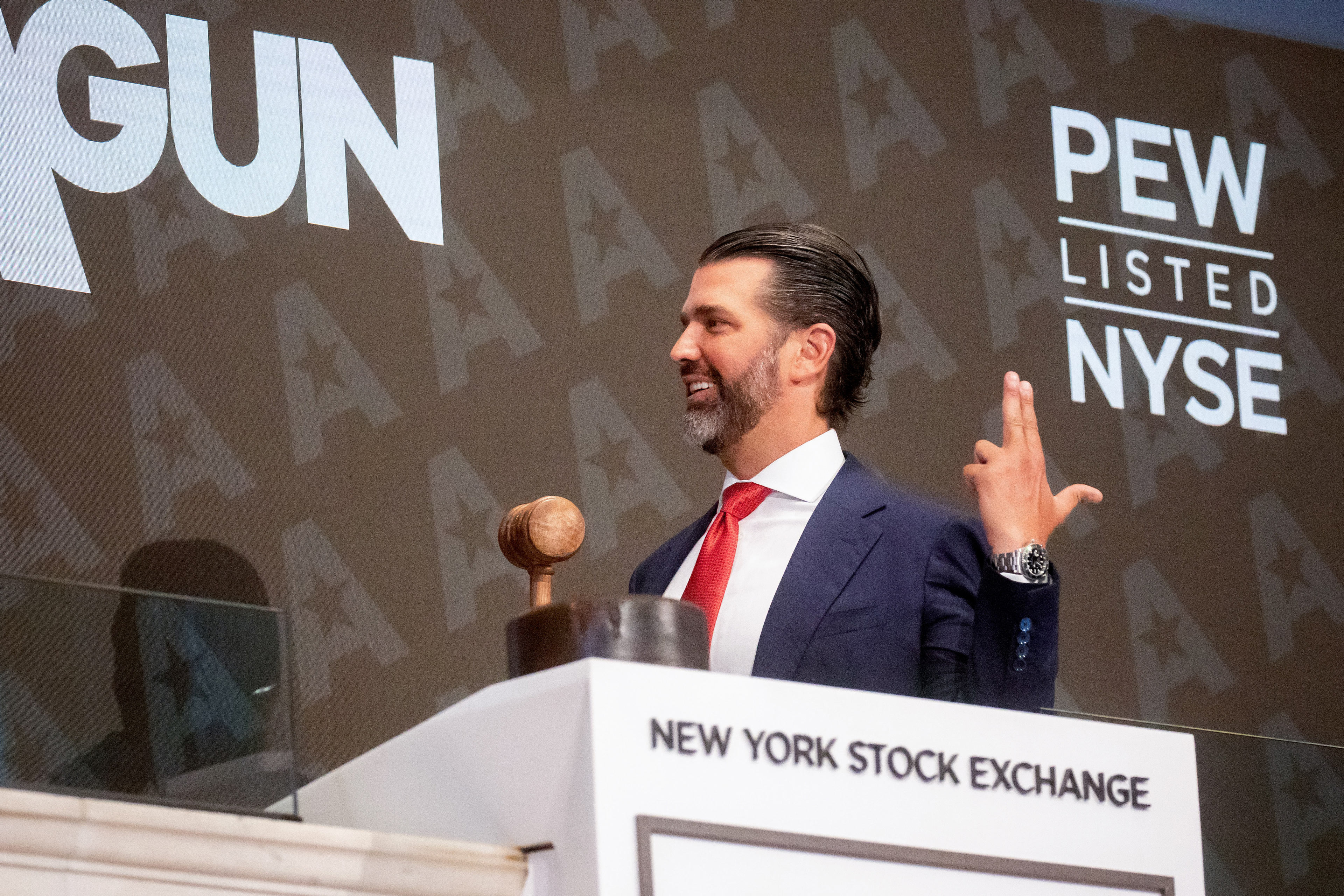 A photo of Donald Trump Jr. at the New York Stock Exchange. He smiles, facing to the left, holding his left hand up in a finger gun pose.
