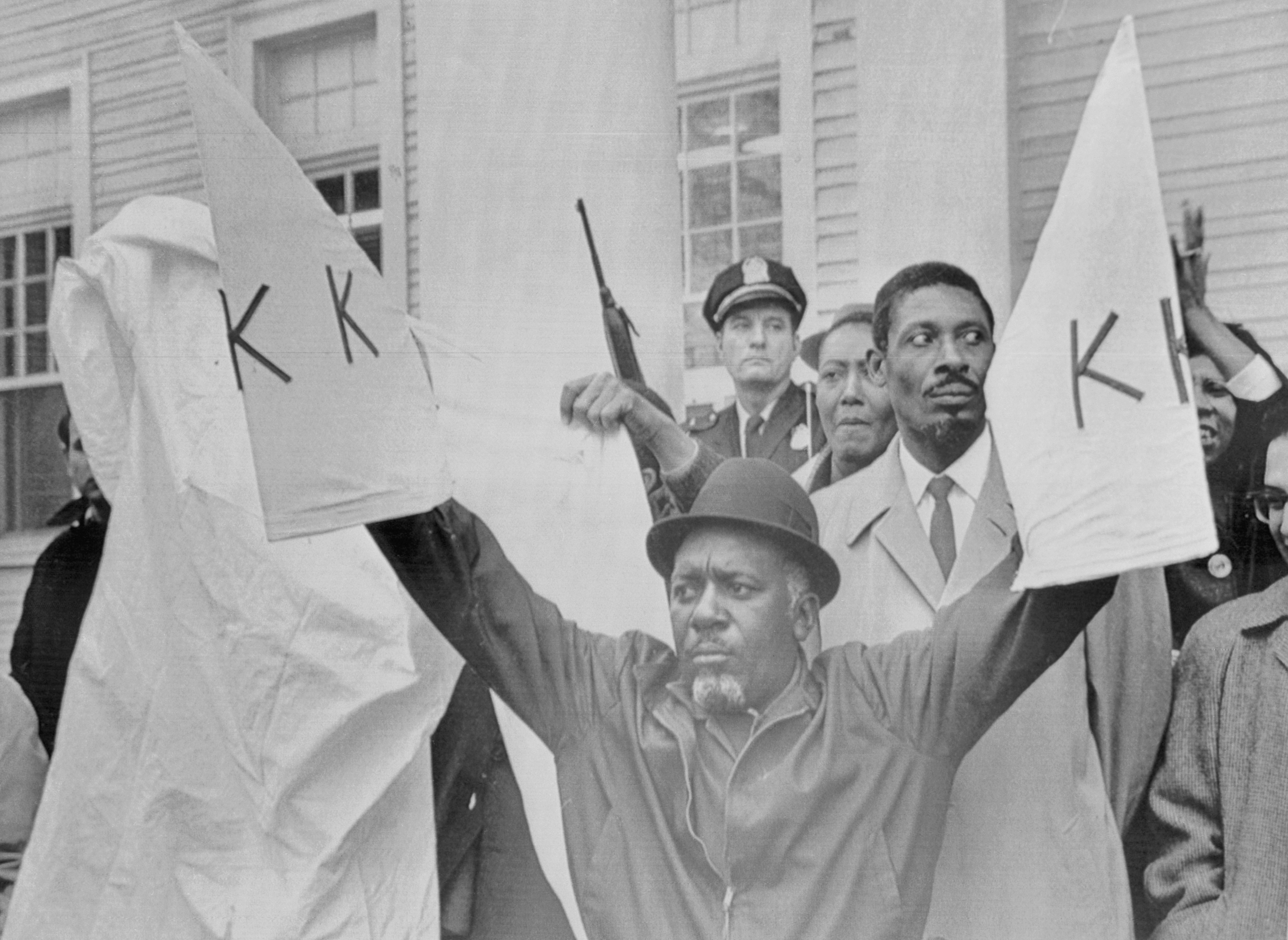 An archival photo of a Black man holding up replica KKK robes at a protest.