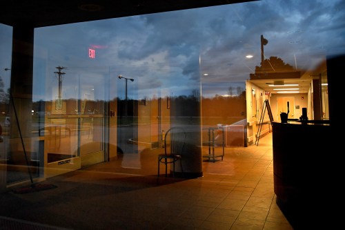A photo of a dimly lit hospital entrance seen through a window. The window is reflecting a view of the parking lot behind the photographer.