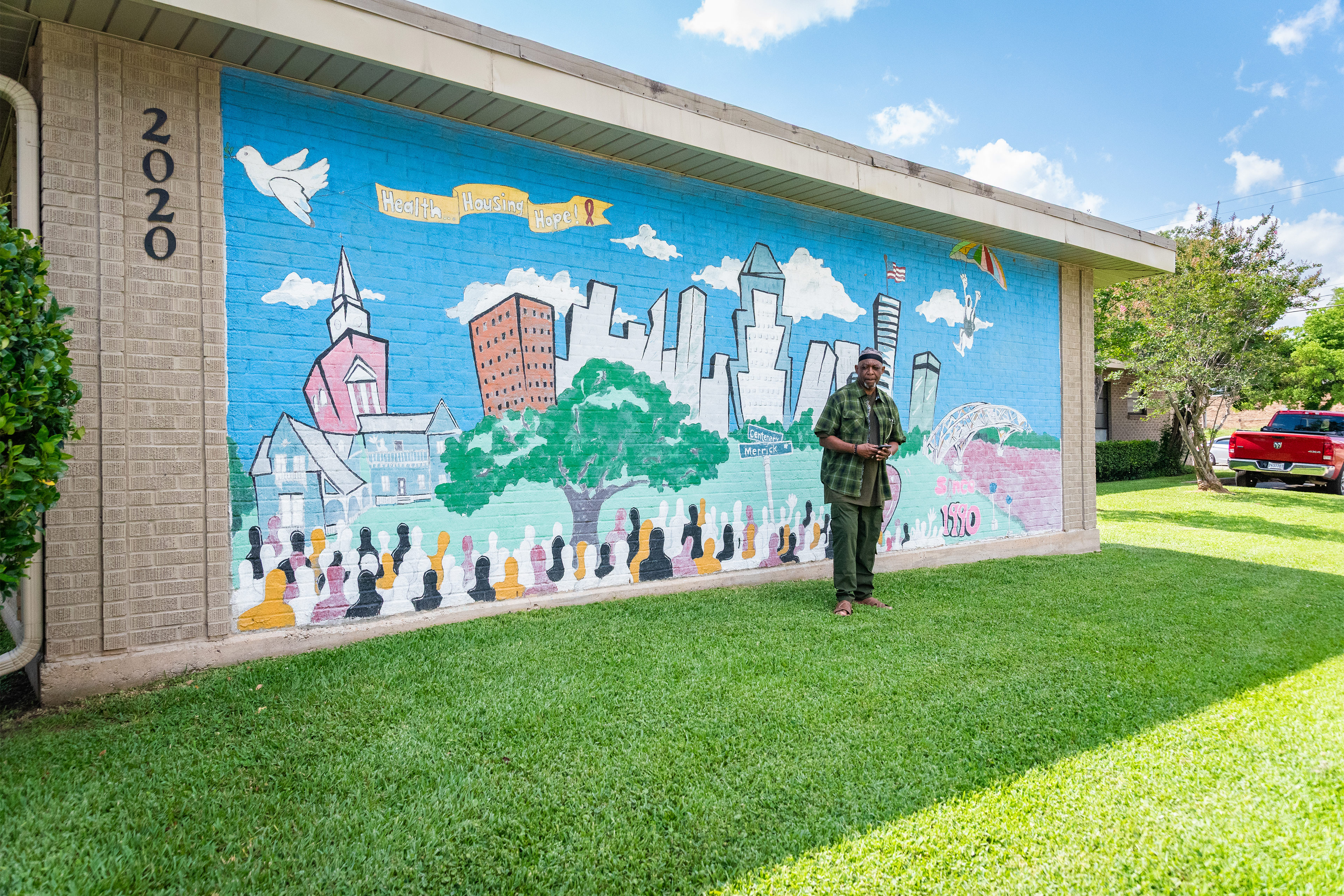 A photo of a man standing outside a community center. A large, colorful mural of  the city is on the façade behind him.