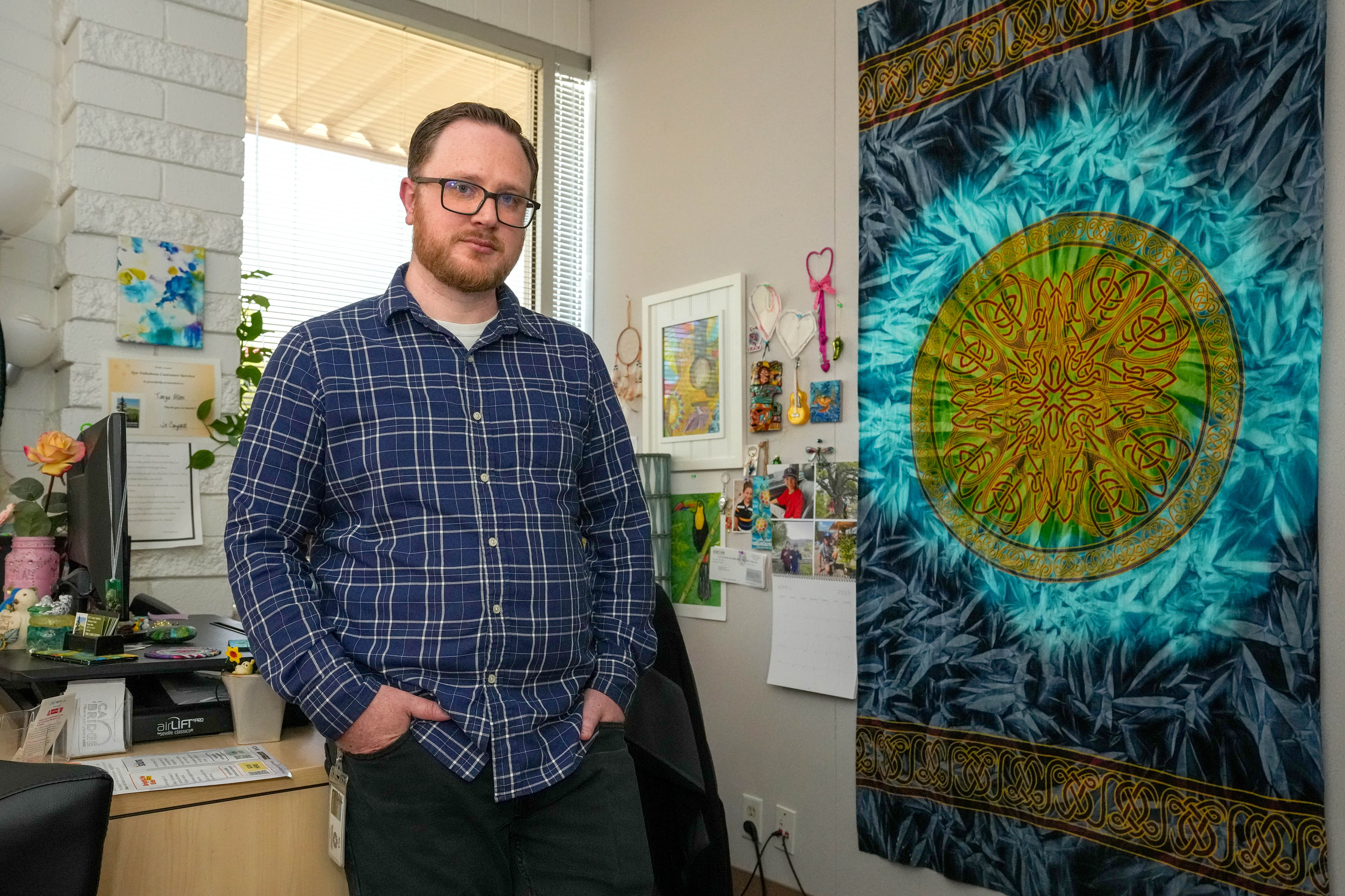 A man wearing a blue plaid button-down shirt and glasses poses for a portrait in an office with a window