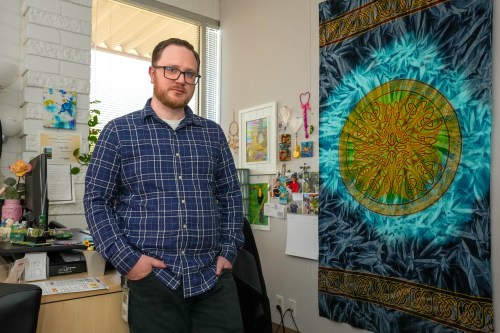 A man wearing a blue plaid button-down shirt and glasses poses for a portrait in an office with a window