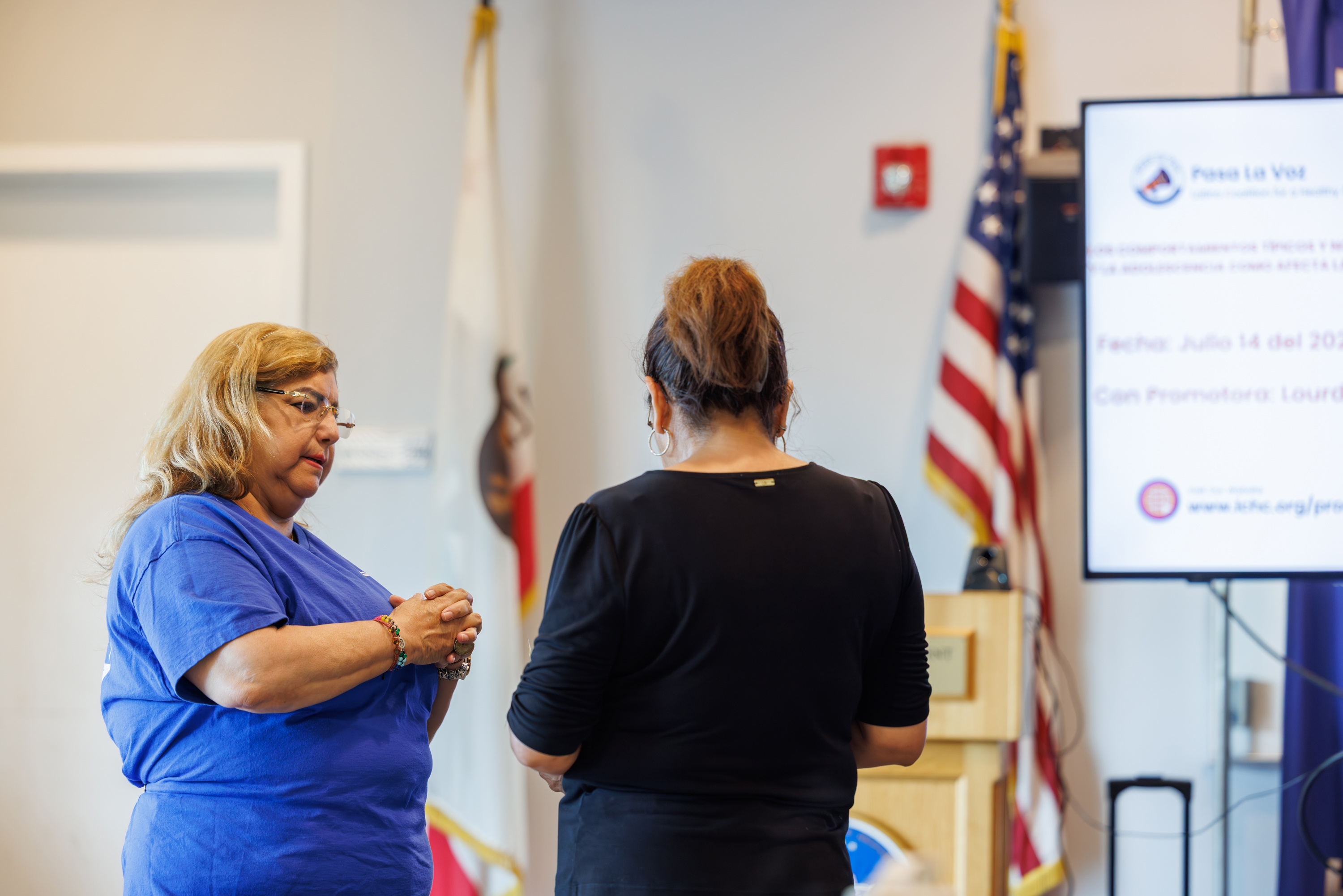 A woman wearing a blue t-shirt and glasses has her hands folded together as she talks with a woman in a black shirt who stands with her back to the camera