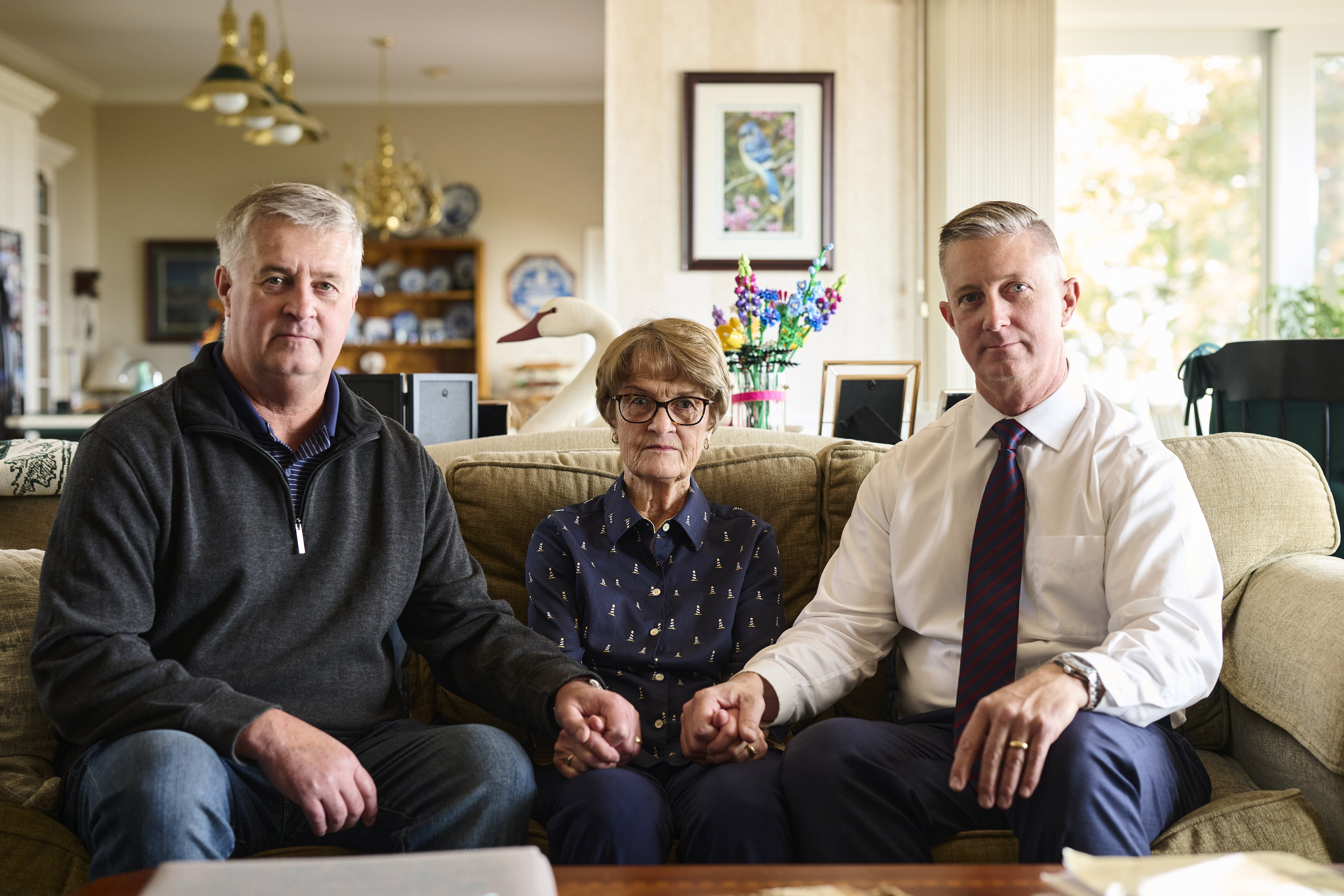 From left, a man, a woman, and another man hold hands while sitting on a couch, posing for a photo