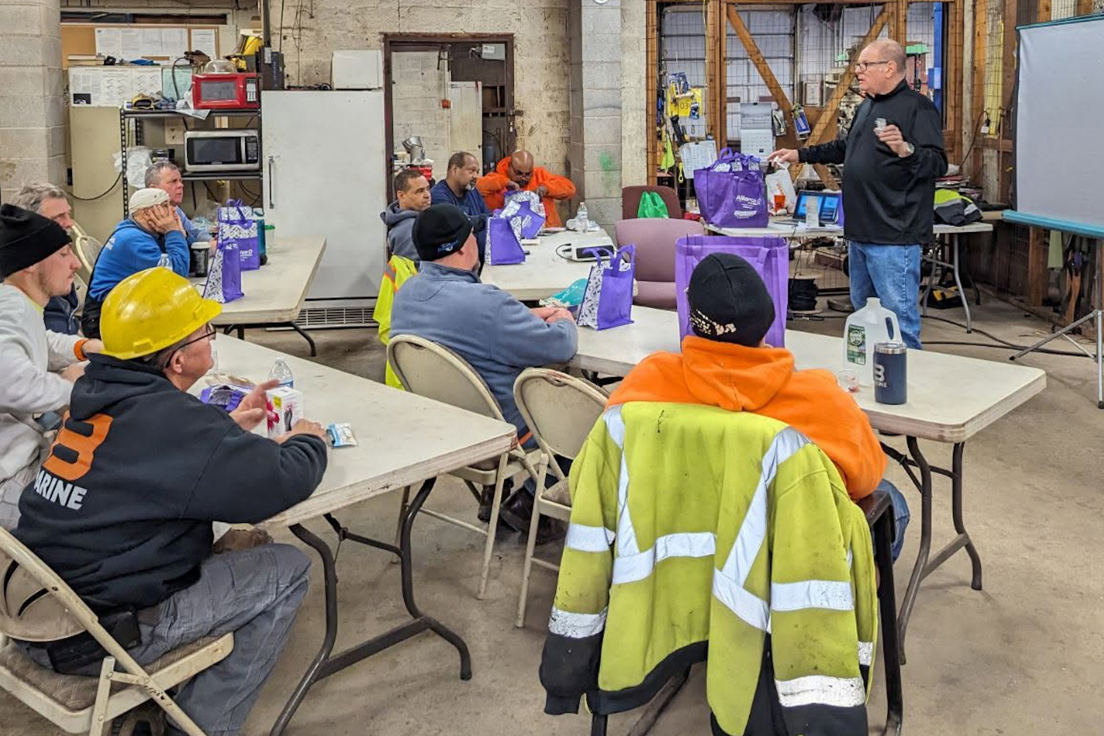A man stands before a room of seated construction workers wearing high-vis clothing and hard hats.