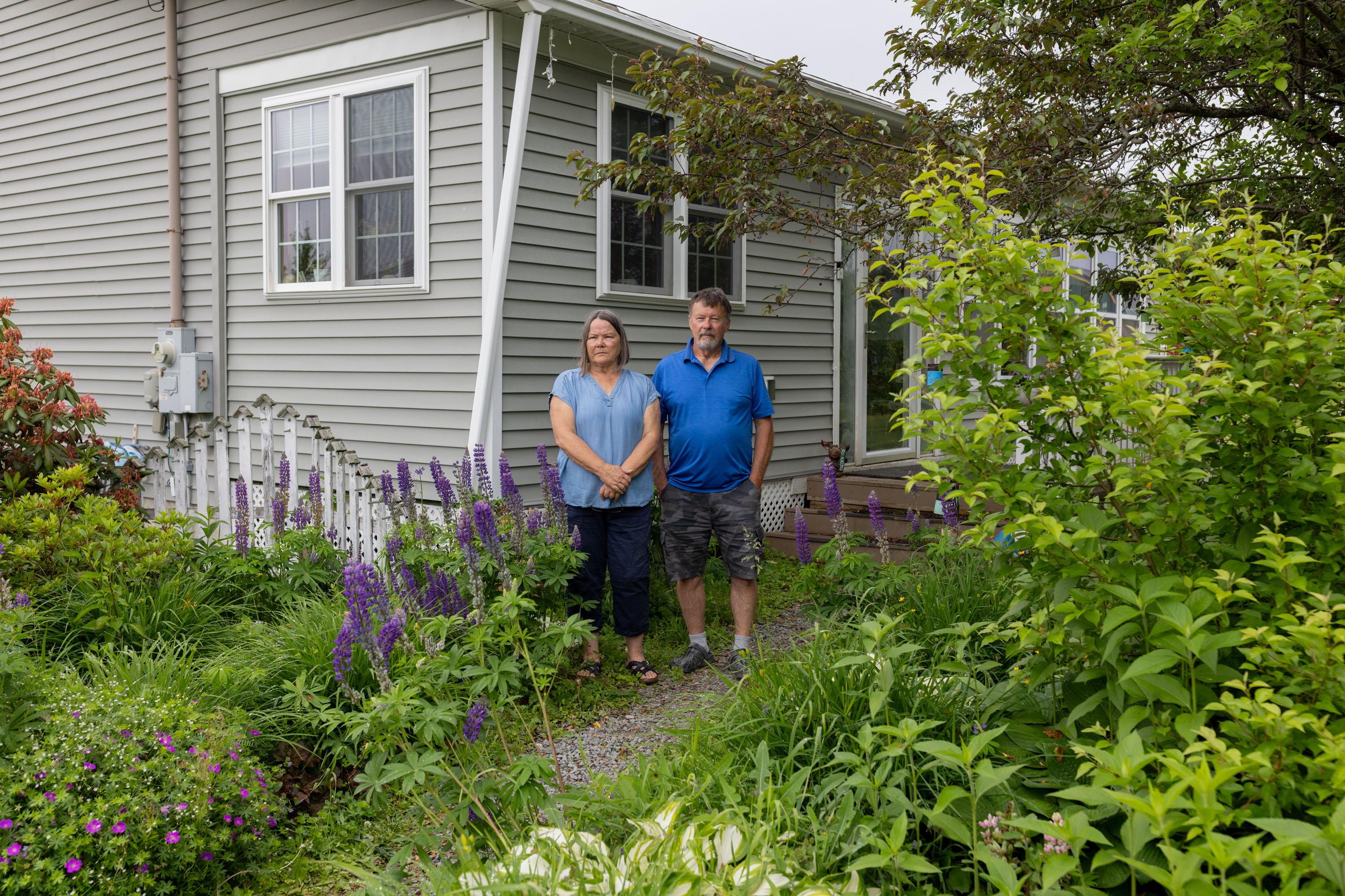 A husband and wife stand outside their home, surrounded by lush green plants and tall purple flowers.