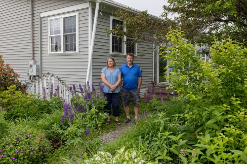 A husband and wife stand outside their home, surrounded by lush green plants and tall purple flowers.