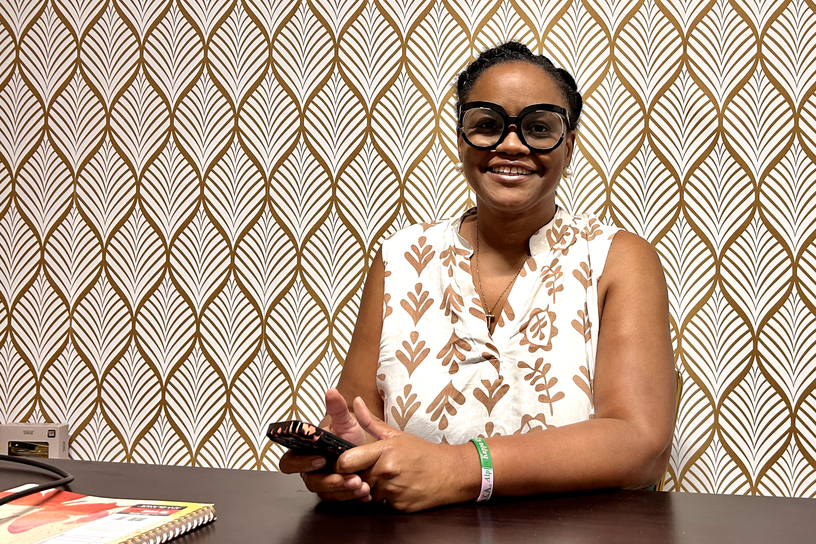A woman wearing black-rimmed glasses and a white-and-brown patterned top sits at a table holding a cell phone while posing for a portrait
