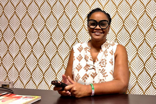 A woman wearing black-rimmed glasses and a white-and-brown patterned top sits at a table holding a cell phone while posing for a portrait