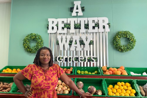 A photo of a Black woman posing for a portrait in the produce section of her grocery store. A sign behind her bears the store's name: A Better Way Grocers.